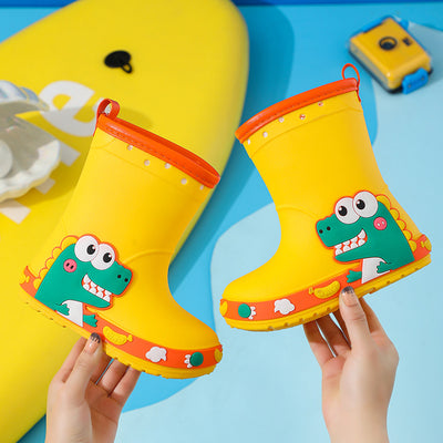 Bright yellow children's rain boots with a green cartoon dinosaur design and orange trim, held by hands. - Image 9