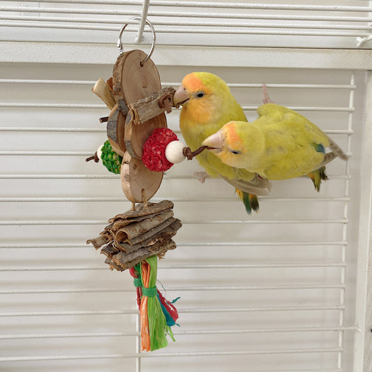 Two yellow-green parrots playing with a hanging bird bite toy made of natural wood slices, bark, and colorful shredded paper.