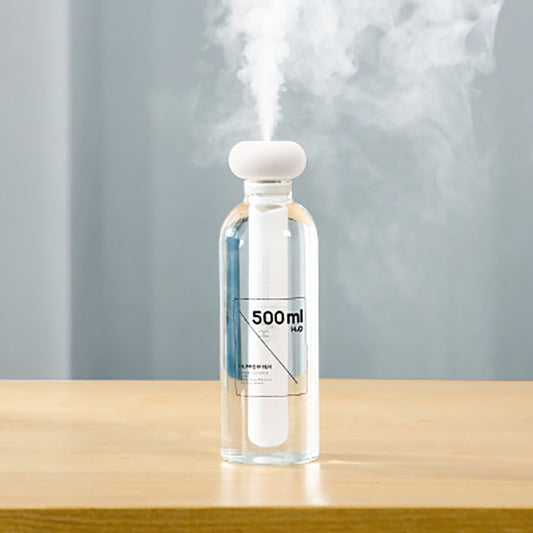 Clear bottle-shaped USB humidifier with a white donut-style top, emitting a stream of cool mist on a wooden table.