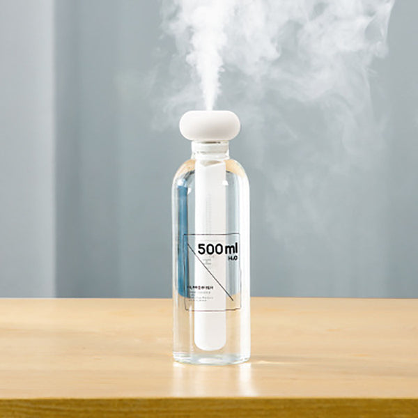 Clear bottle-shaped USB humidifier with a white donut-style top, emitting a stream of cool mist on a wooden table.