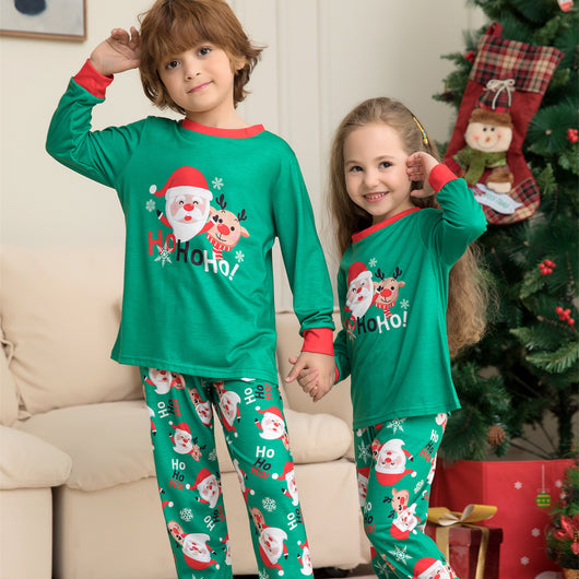 Family of four wearing matching green Christmas pajamas with Santa Claus and reindeer print, sitting on a sofa.